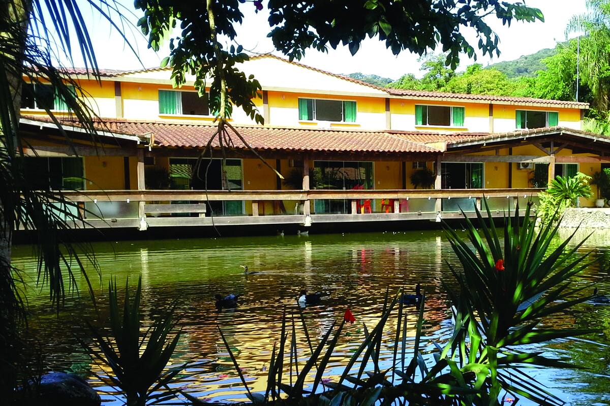 Um edifício com fachada amarela e telhado de telhas vermelhas, um lago com nenúfares e uma ponte.
