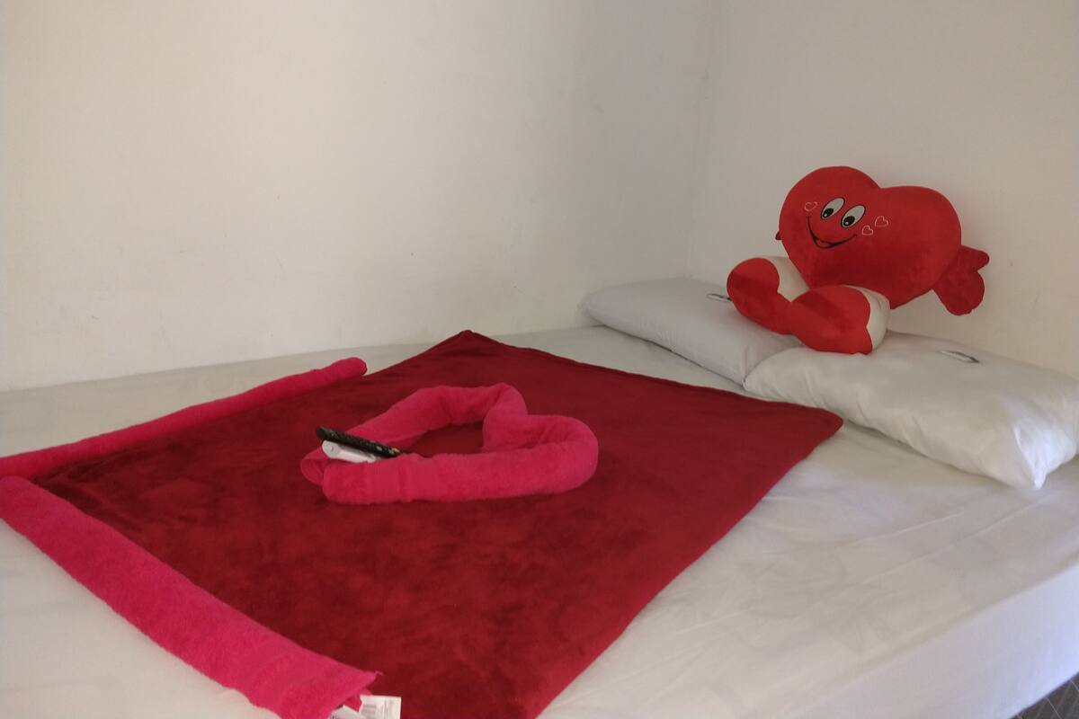 Uma cama com um cobertor vermelho, um brinquedo de pelúcia vermelho e uma almofada branca.