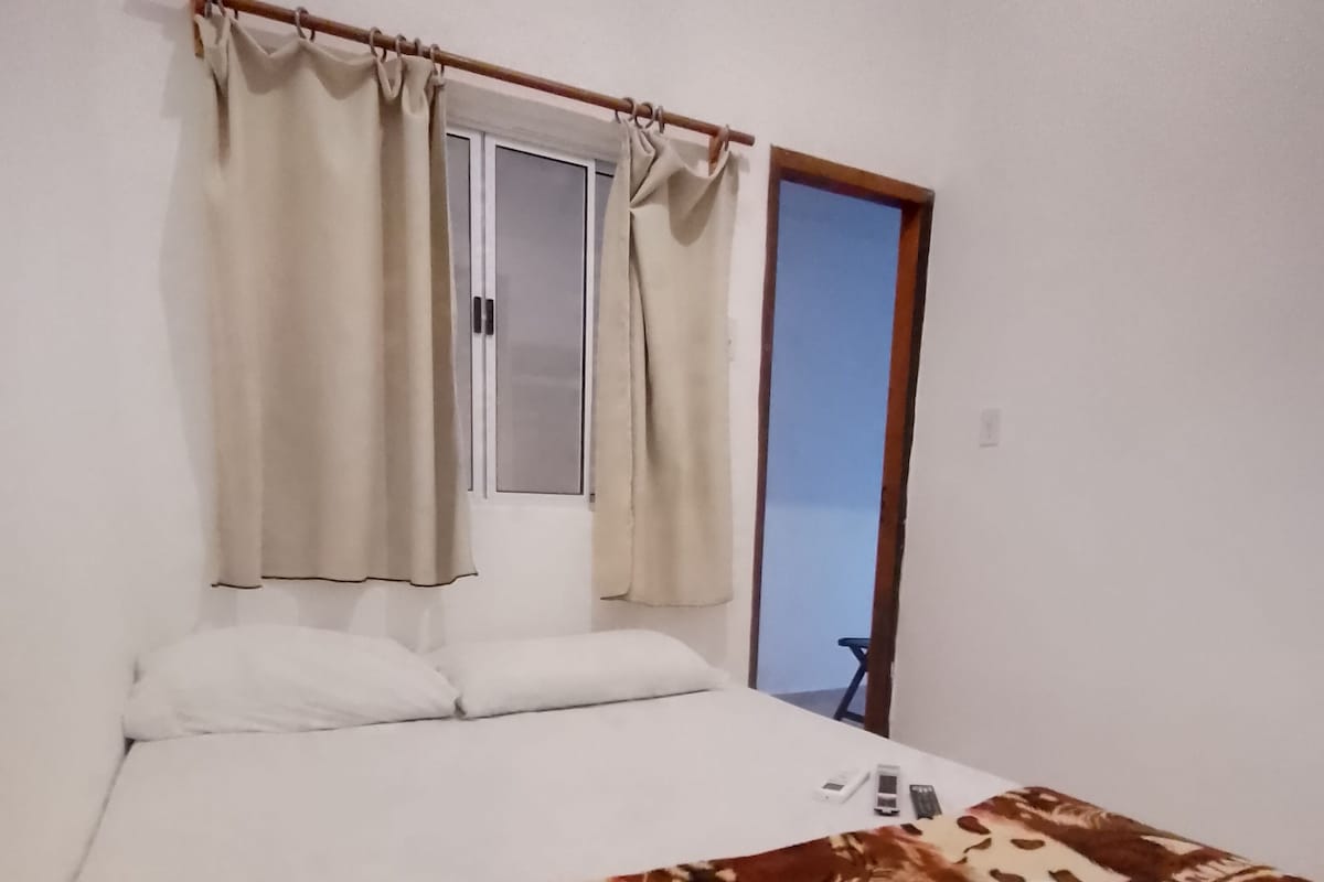 Un dormitorio con cama, ventana con cortinas, puerta y vista al exterior.
