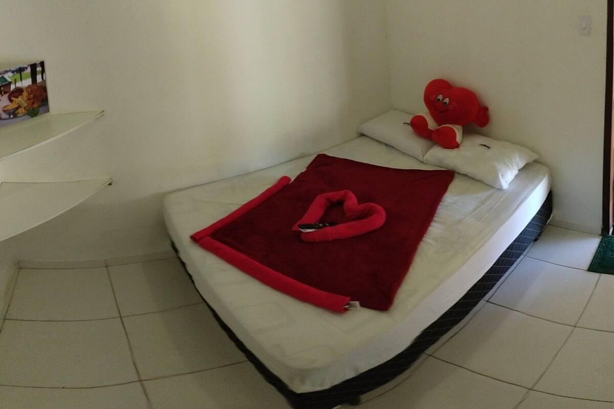 Uma cama com travesseiro e cobertor em formato de coração, uma pequena prateleira com uma foto, e um tapete verde na entrada.