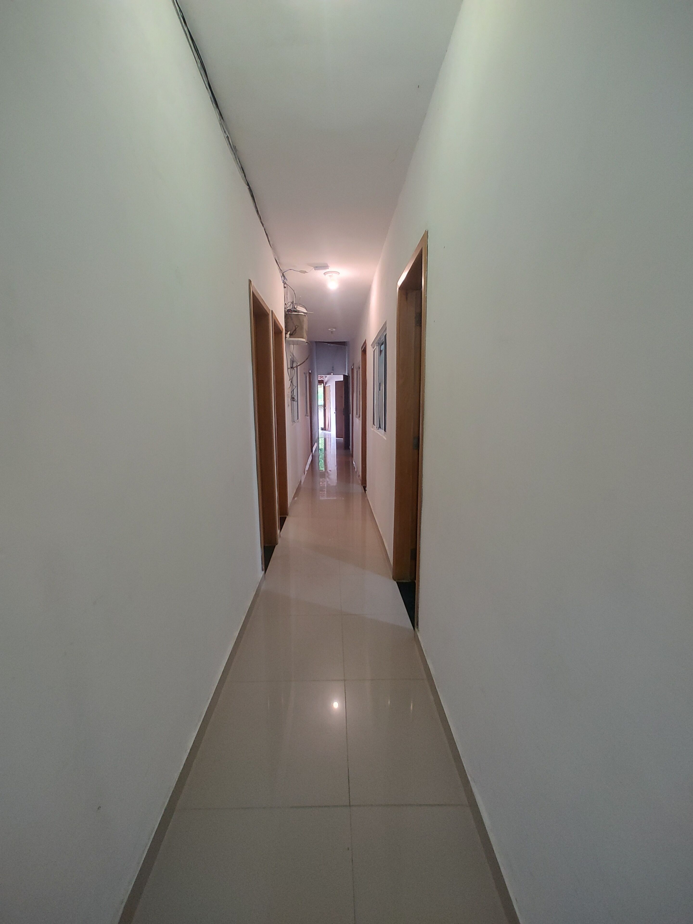 Un passadís llarg i estret amb parets blanques i portes de fusta, que condueix a una habitació lluminosa al final.