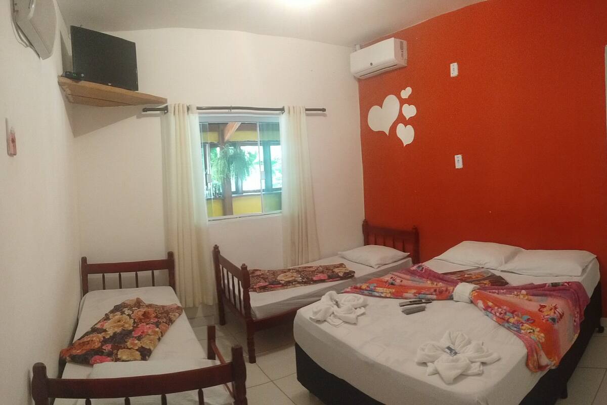 Quarto com duas camas, janela com cortinas, ar condicionado fixado na parede e televisão em uma estante.
