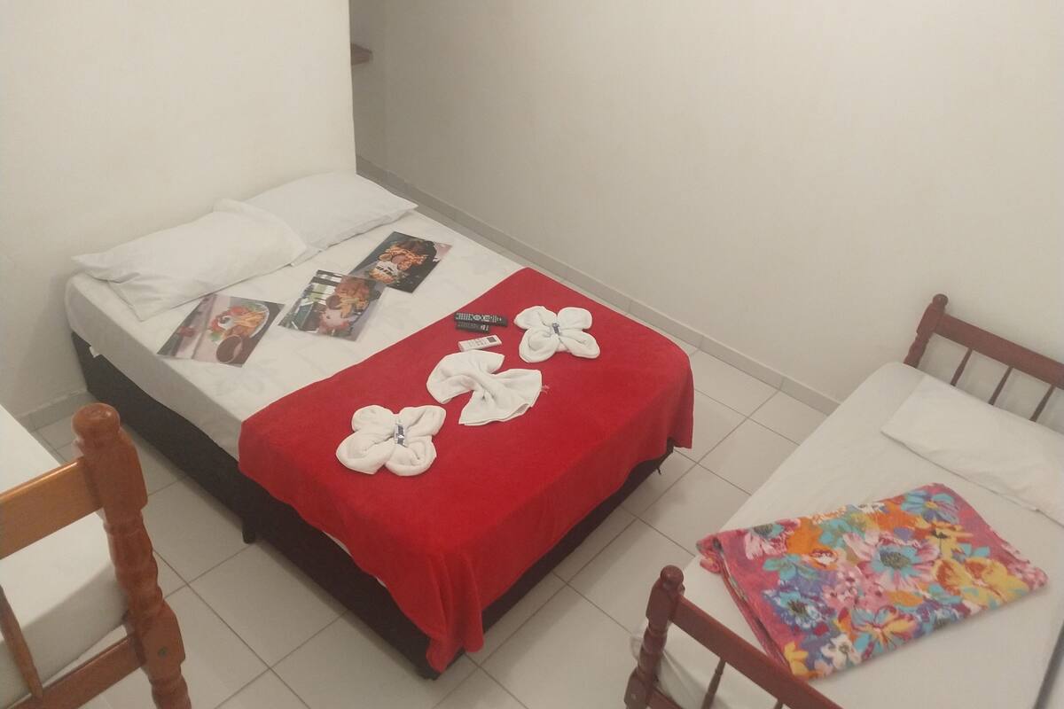 Uma cama pequena e bem arrumada com um cobertor vermelho e almofadas brancas, uma cadeira de madeira e uma mesa com toalha de mesa florida.