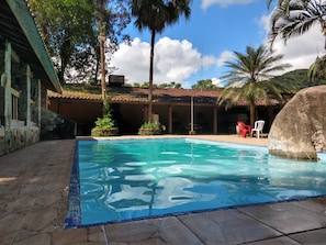 A swimming pool with clear water, surrounded by stone tiles, palm trees, and a building with a tiled roof. - Rancho Texas Ubatuba (Ubatuba)