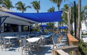 An outdoor seating area with tables and chairs, shaded by blue canopies, and surrounded by palm trees.