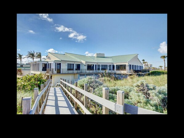 A beachfront property with a wooden boardwalk leading to a house with a green roof and multiple balconies.