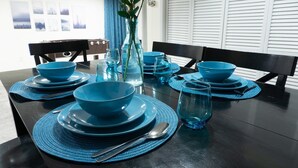 Een eettafel gedekt met blauw servies en placemats, een vaas met bloemen, en een raam met jaloezieën op de achtergrond.