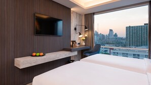 A modern hotel room with a large bed, a mounted TV, a desk with a chair, a small shelf with fruits, and a city view through large windows.