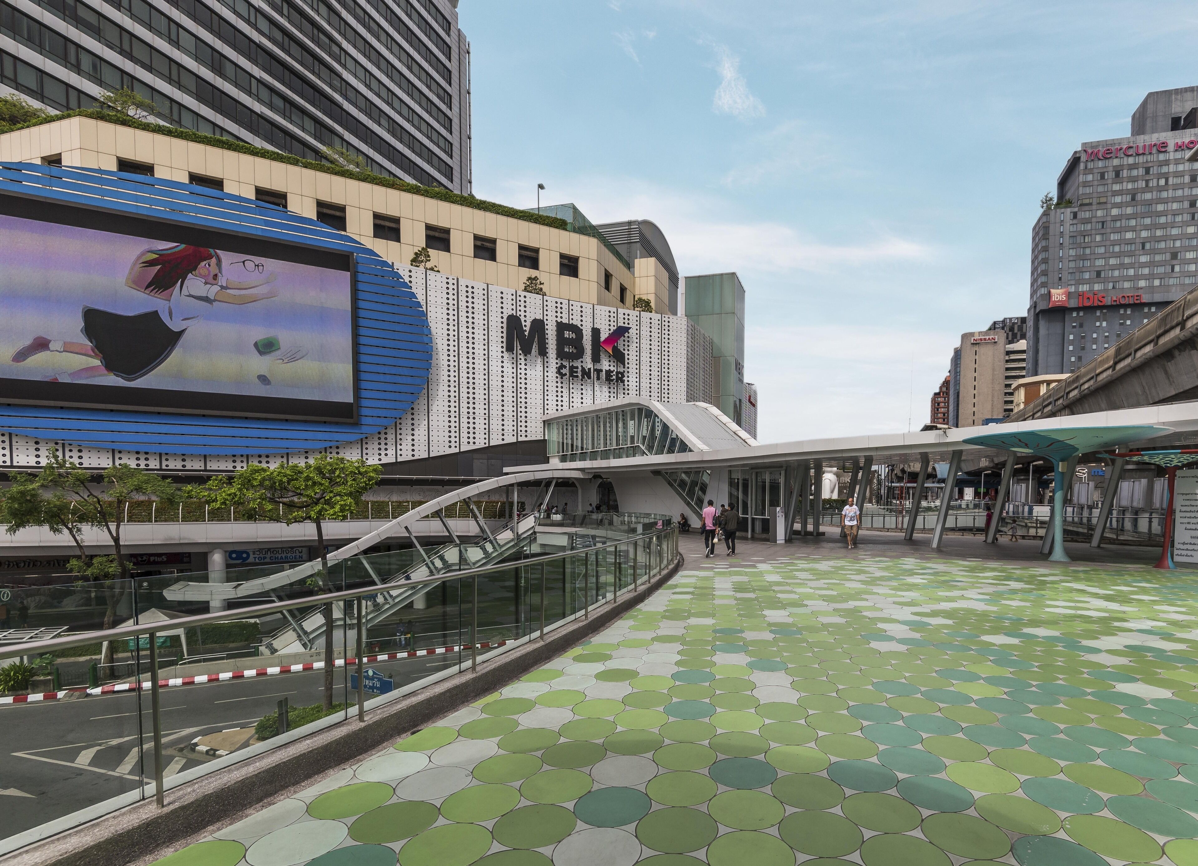 Un edificio moderno del centro comercial MBK con un gran cartel digital, una zona peatonal cubierta y un suelo con diseño verde.