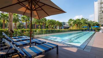 A swimming pool area with lounge chairs and umbrellas, surrounded by palm trees and a cityscape in the background.
