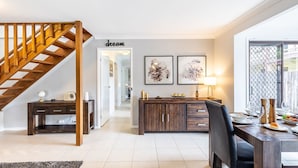 A modern living room with a wooden staircase, a dining area with a table set for two, and a kitchen visible in the background. - Willows At The Bay, 111 Horace St - Large House with Aircon, WIFI and Boat Parking (Shoal Bay)