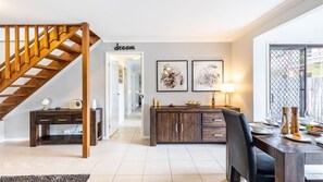 Un salon moderne avec un escalier en bois, un coin repas avec une table dressée pour deux personnes, et une cuisine visible au fond.