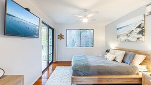 A bedroom with a large bed, a ceiling fan, a view of the sea through a window, and a beach scene painting. - Willows At The Bay, 111 Horace St - Large House with Aircon, WIFI and Boat Parking (Shoal Bay)