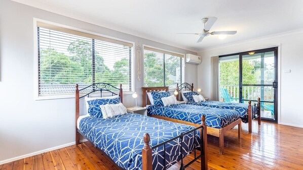 A bedroom with two beds, large windows, a ceiling fan, and a view of greenery outside.