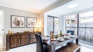 A dining area with a wooden table set for six, a wooden cabinet with drawers, and two framed floral artworks on the wall. - Willows At The Bay, 111 Horace St - Large House with Aircon, WIFI and Boat Parking (Shoal Bay)