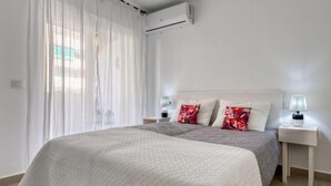 A hotel room with a large bed, white curtains, a white headboard, two bedside tables with lamps, and an air conditioning unit on the wall. - Urbatenis by Interhome (Jávea)