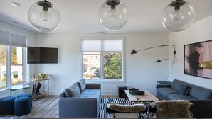 A modern living room with a large window, a flat-screen TV, a sofa, ottomans, and a coffee table. - Piedmont Place 304 | Walkable to Downtown Crozet (Crozet)