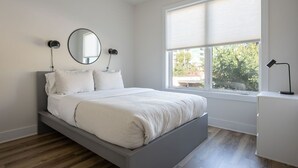 A modern bedroom with a bed, two bedside lamps, a round mirror, and a window with blinds. - Piedmont Place 304 | Walkable to Downtown Crozet (Crozet)