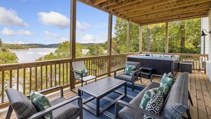 A covered outdoor deck with seating, a hot tub, and a view of a lake and forest. - The Dogwood - Hot Tub - Lake Front Access (Springdale)