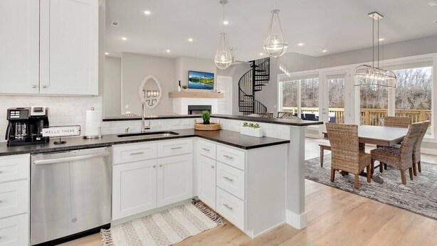 A modern kitchen with white cabinetry, a central island, and a dining area with wicker chairs. - The Dogwood - Hot Tub - Lake Front Access (Springdale)