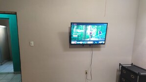 Una stanza con una televisione a parete, un letto con copriletto blu, un comò e un pavimento piastrellato.