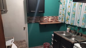 Una cucina compatta con lavello, fornello e pensili. Un piccolo cestino è vicino alla porta.