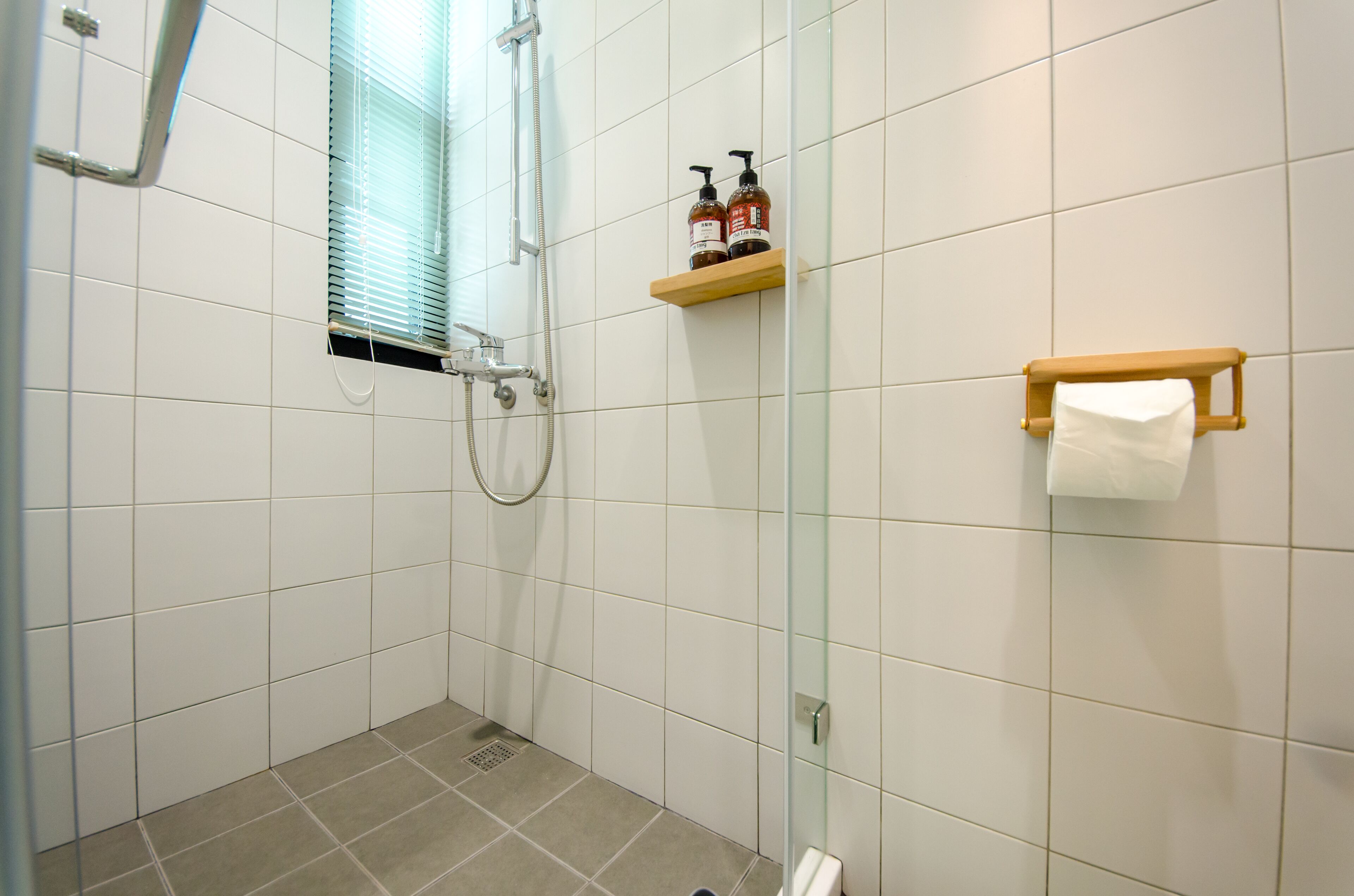 Un espacio de ducha pequeño con azulejos, una puerta de cristal, un estante de madera con artículos de aseo y un perchero para toallas.