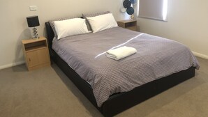 A neatly made bed with a patterned bedspread, two white pillows, a folded white towel, a bedside lamp, and a small wooden nightstand with a lamp. - JoyWaves New  4Bedroom retreat - ultimate getaway
 (Kambah)