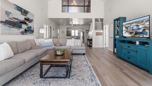 A spacious living room with a high ceiling, a large painting, a sofa set, a coffee table, and a television on a blue cabinet. - Private Heated Pool, walkout on the beach! - Island Star (Dauphin Island)