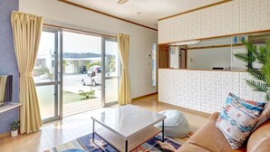 A modern living room with a large sliding glass door, a ceiling fan, a white brick wall, and a leather sofa with patterned cushions. - Kailana Villa (Yomitan)