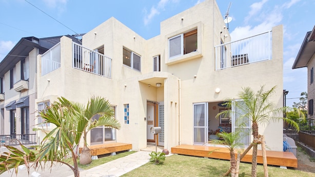 A modern two-story building with a flat roof, large windows, and a small front yard with palm trees and a bench. - Kailana Villa (Yomitan)