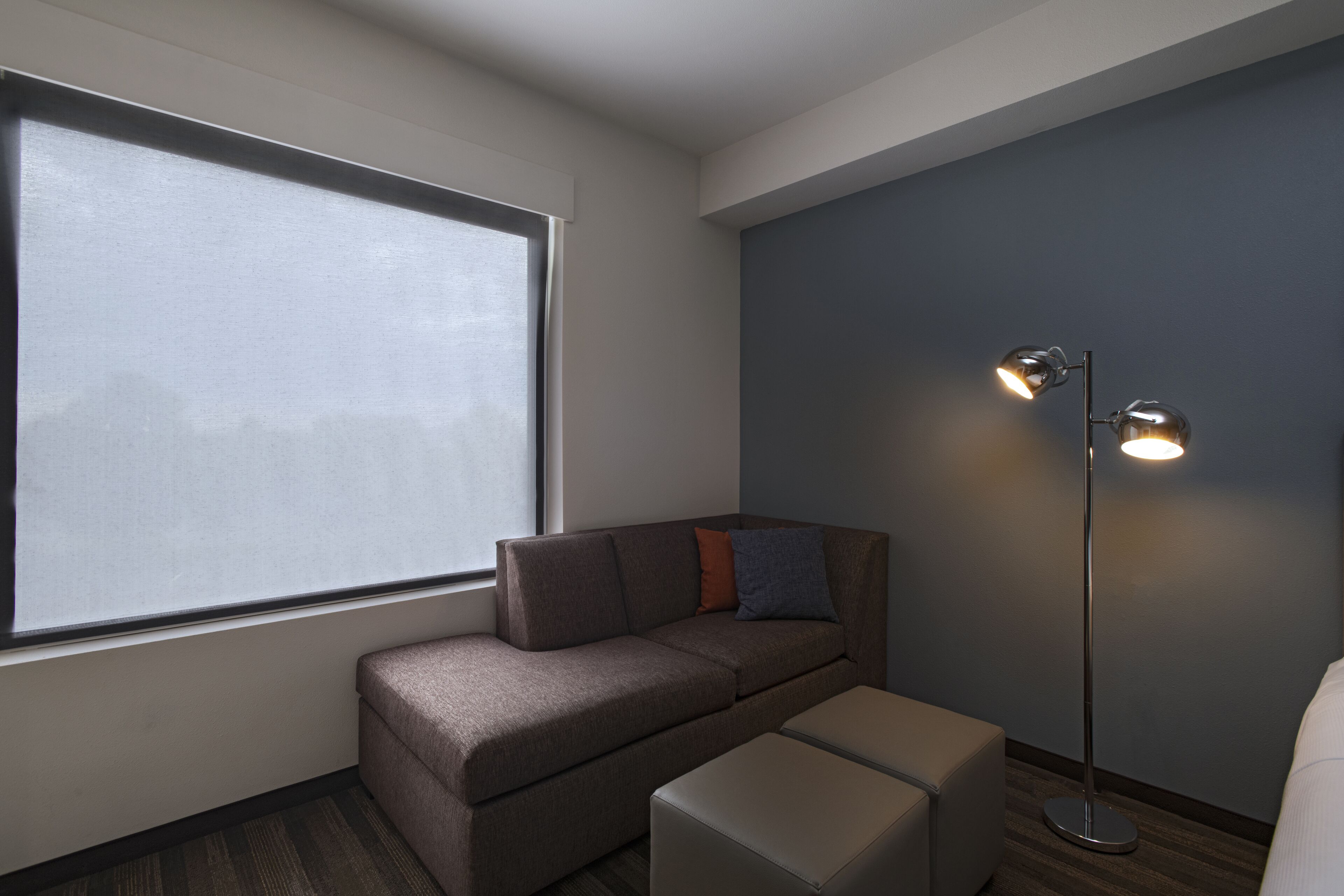 A modern hotel room with a large window, a sofa, a floor lamp, and a small ottoman.