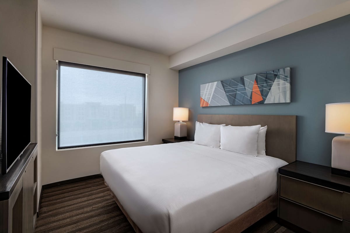 A hotel room with a large bed, two bedside tables with lamps, a flat-screen TV, and a window with blinds.