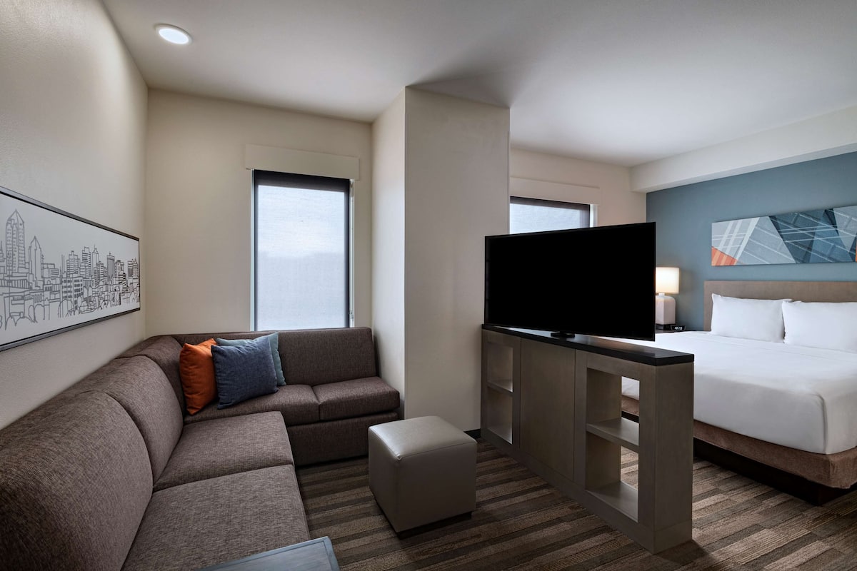 A hotel room with a sofa, ottoman, flat-screen TV, and a bed with white bedding.