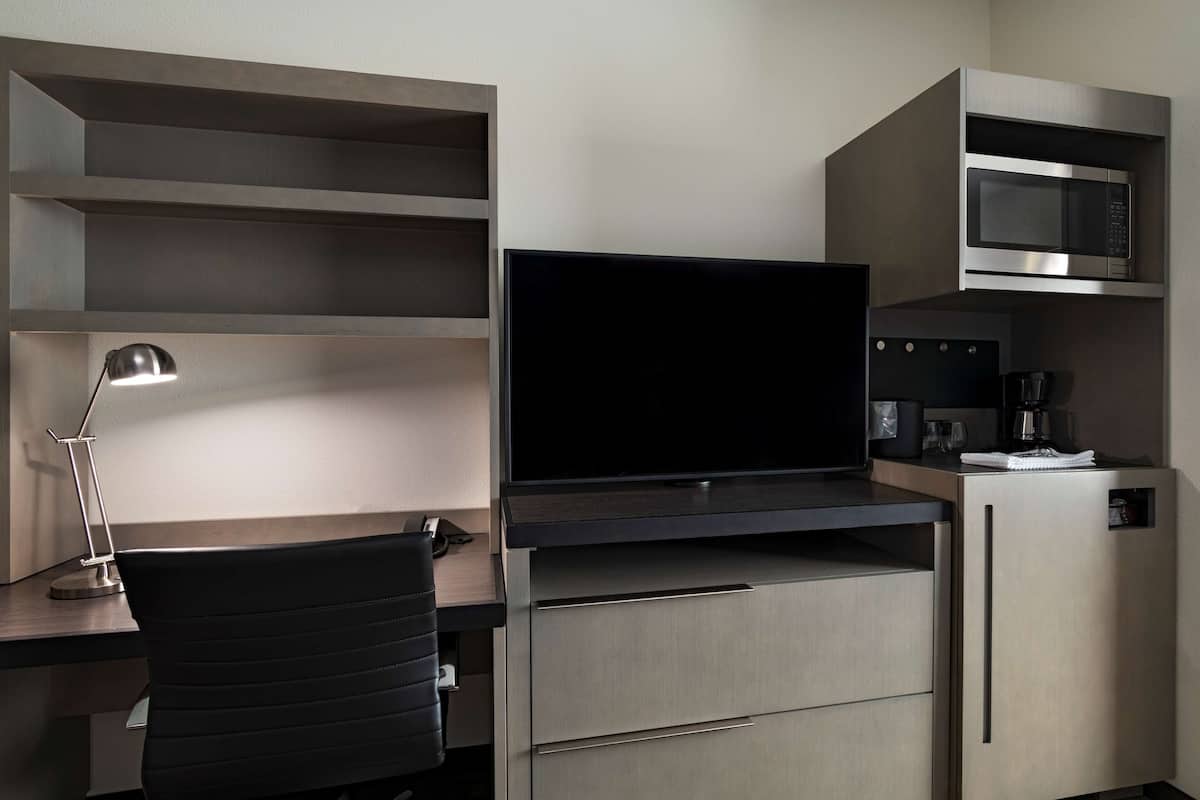 A modern hotel room with a flat-screen TV, a desk with a lamp, a microwave, and a coffee machine.