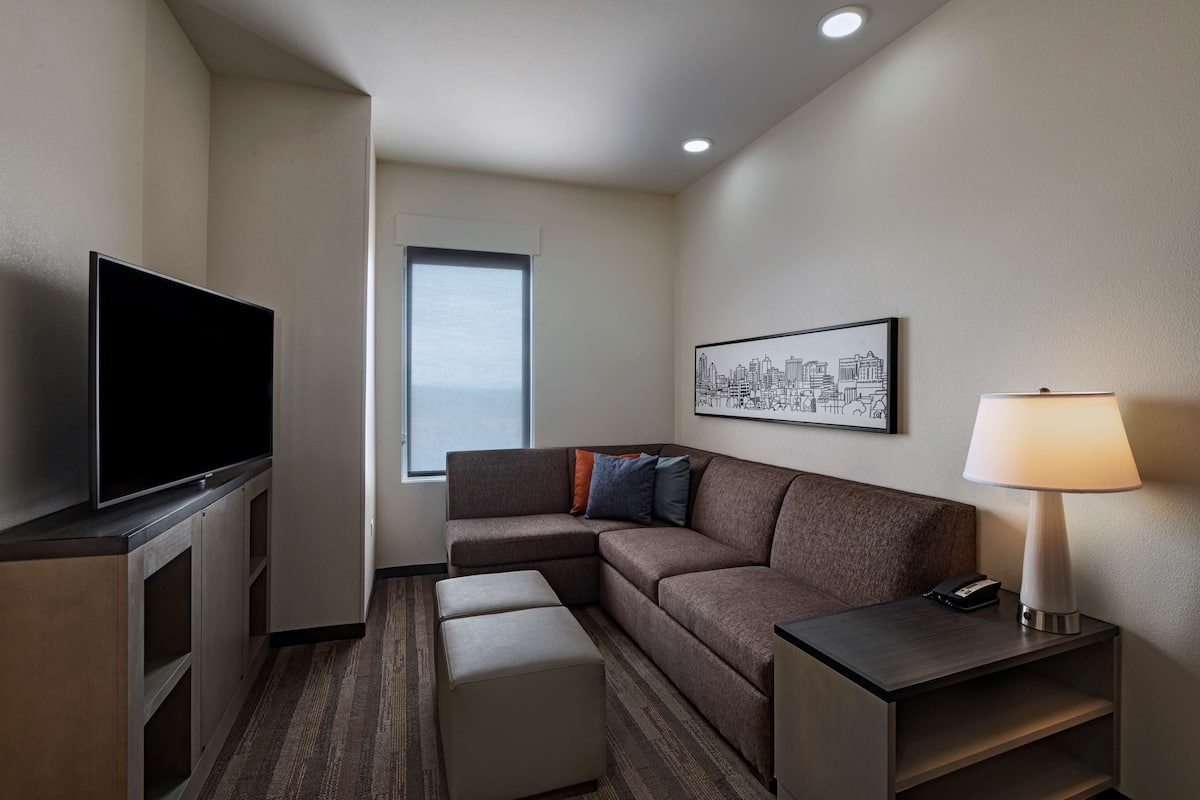 A hotel room with a flat-screen TV, a sofa, a small ottoman, a nightstand with a lamp, and a cityscape artwork on the wall.