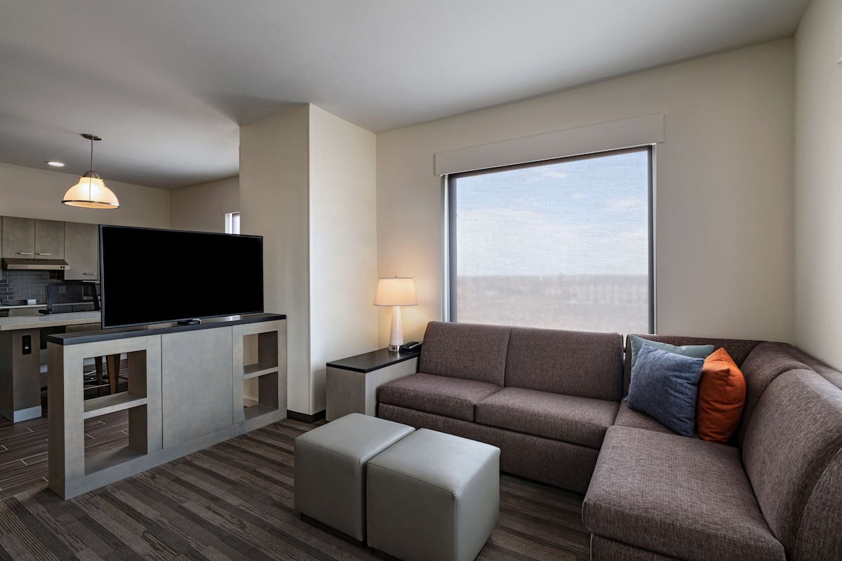 A modern hotel room with a flat-screen TV, a sofa, a lamp, and a large window offering a city view.