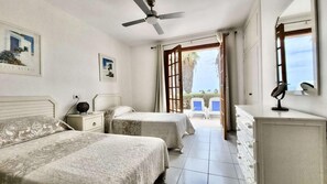 Ein Schlafzimmer mit zwei Betten, einem Deckenventilator, einem Schminktisch und Blick ins Freie.