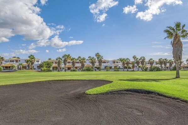 A golf course with a sand bunker, palm trees, and a row of villas in the background.