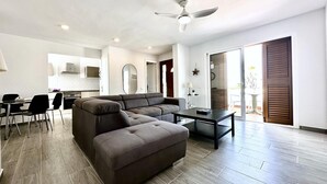 A modern living room with a sectional sofa, a dining area with chairs, a kitchen in the background, and a sliding glass door leading to an outdoor area.