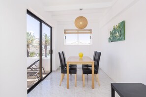 A dining area with a wooden table and four chairs, a hanging light fixture, a window with blinds, and a painting on the wall.