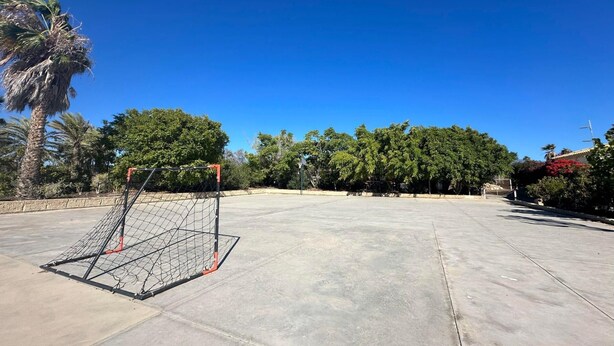 An outdoor basketball court with a goal post, surrounded by trees and a clear blue sky.