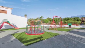 A playground with swings and a slide, surrounded by green grass and a white building.