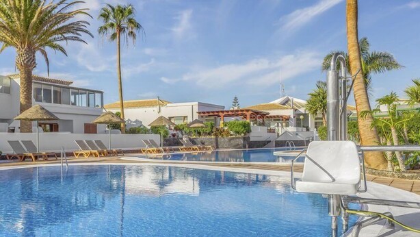 En swimmingpool med liggestole og parasoller, omgivet af palmer og bygninger.