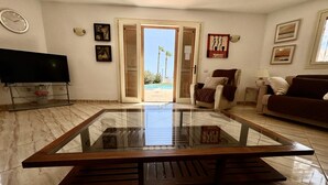 Un salon spacieux avec une table basse en verre, un téléviseur à écran plat et une vue sur la plage, visible à travers la porte ouverte.