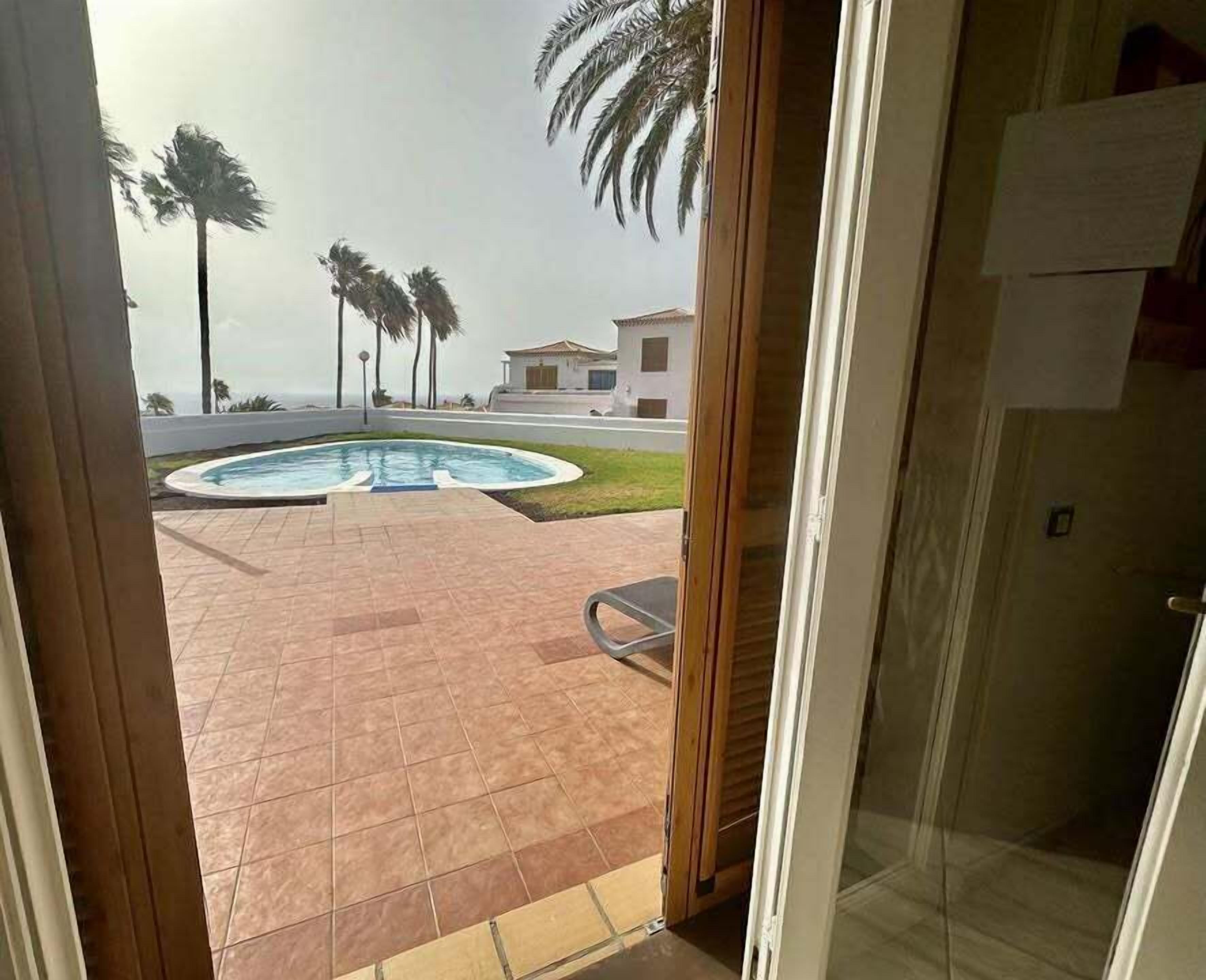 A view from a room to an outdoor swimming pool area with palm trees and a building in the background.