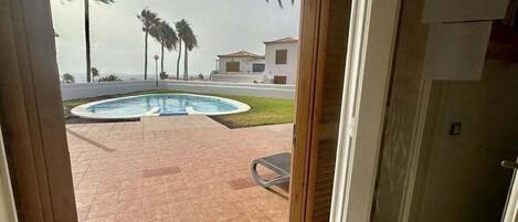 A view from a room to an outdoor swimming pool area with palm trees and a building in the background.