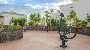 Un espace extérieur aménagé pour faire de l’exercice, équipé de divers appareils de musculation, entouré d’un jardin paysager avec des palmiers et un bâtiment blanc en arrière-plan.
