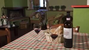 A dining table with a checkered tablecloth, two wine glasses, a bottle of wine, a basket of bread, and a wicker chair.