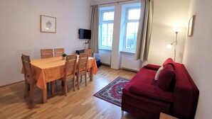 Ein Zimmer mit Holztisch, Stühlen, einer roten Couch, einem an der Wand befestigten Fernseher und mit Vorhängen versehenen Fenstern.
